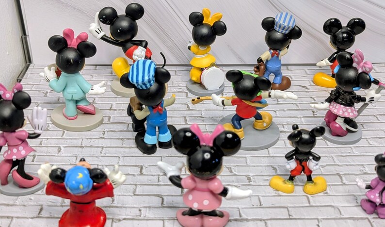 May include: A collection of Mickey Mouse figurines in various poses and outfits. The figurines are black with white gloves and shoes, and some have colorful clothing and accessories. Some are playing musical instruments.