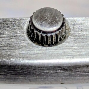 Puede incluir: Primer plano de una corona de reloj, un componente circular peque&ntilde;o y texturizado. La corona est&aacute; hecha de un material met&aacute;lico opaco y est&aacute; rodeada por una superficie met&aacute;lica cepillada. El borde de la corona tiene un dise&ntilde;o estriado.