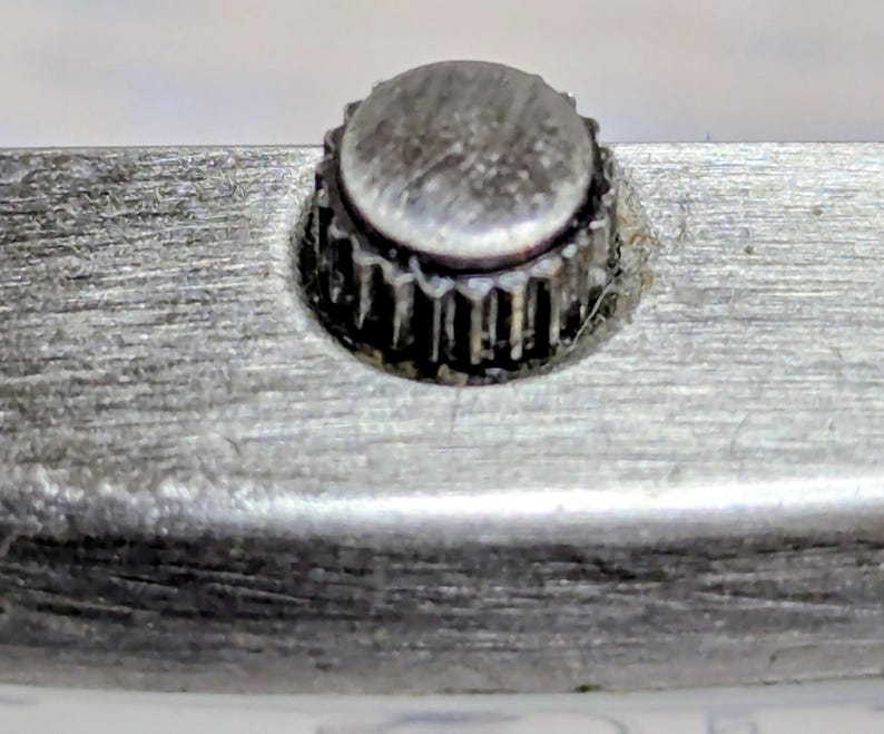 Puede incluir: Primer plano de una corona de reloj, un componente circular peque&ntilde;o y texturizado. La corona est&aacute; hecha de un material met&aacute;lico opaco y est&aacute; rodeada por una superficie met&aacute;lica cepillada. El borde de la corona tiene un dise&ntilde;o estriado.