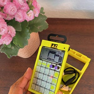 May include: A yellow Pocket Operator electronic music device with a screen and buttons. The device has a black handle and the words "pocket operator" printed on it. A yellow case and a black audio cable are also visible.
