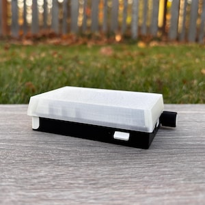 May include: A rectangular black and white device with a white, textured lid. A small white button and a black cylindrical component are visible on the side. The device is resting on a gray wooden surface.