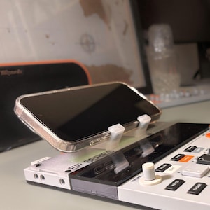 Pode incluir: Um suporte de plástico branco impresso em 3D segura um smartphone numa capa transparente, posicionado sobre um dispositivo de produção musical eletrónico branco e preto com a etiqueta EP SERIES, botões, um botão de volume e um ecrã preto.