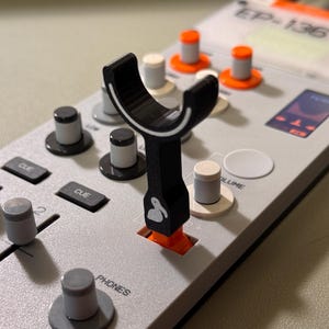 Peut inclure: Un capuchon de fader noir en forme de Y avec une icône de pélican blanche fixé sur un curseur orange d'un appareil de production musicale EP-136 gris clair. L'appareil comporte plusieurs boutons gris et orange, des touches CUE et VOLUME.
