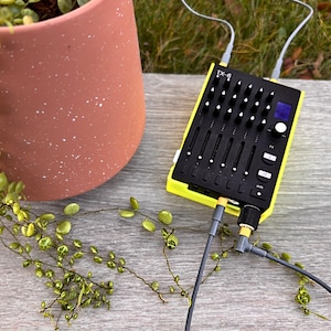 May include: A black and yellow audio mixer with multiple sliders, knobs, and ports. The mixer is connected to cables and sits on a wooden surface next to a terracotta pot with a trailing plant. The mixer has the text "TX-6" on it.