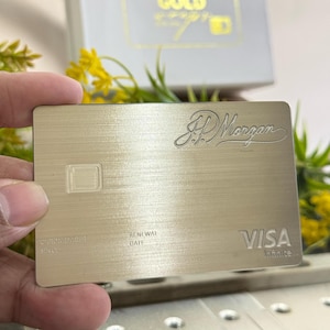 Op de afbeelding: Een goudkleurige Visa Infinite creditcard met het JP Morgan logo. De kaart heeft een geborstelde metalen afwerking en het woord "VISA" is zichtbaar. Een witte doos met het woord "GOLD" staat op de achtergrond.
