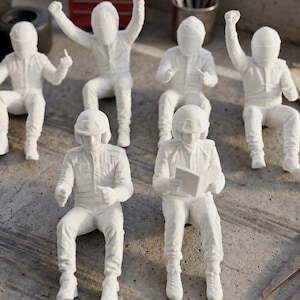 May include: A collection of seven white miniature racing driver figurines in various poses, including standing, sitting, and cheering. The set includes black tires, a red toolbox, a metal cup with tools, and a metal caliper tool on the ground.