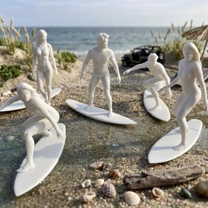 Könnte beinhalten: Ein Set aus fünf weißen Miniaturfiguren, die Personen auf Surfbrettern darstellen. Die Figuren nehmen verschiedene Surf-Posen auf weißen Surfbrettern ein. Die Szene umfasst ein kleines dunkles Fahrzeug und einen Miniatur-Strohschirm am Sandstrand.