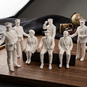 Peut inclure: Un ensemble de sept figurines miniatures blanches représentant des personnes en tenue de ville formelle, dont plusieurs portent des casquettes à visière. Le groupe comprend des personnages debout et assis sur une surface en bois.