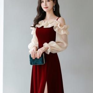 May include: A burgundy velvet evening gown with a high slit, off-the-shoulder design, and sheer, ruffled sleeves. The dress features a sweetheart neckline and is paired with a small, teal clutch. The model is wearing drop earrings.