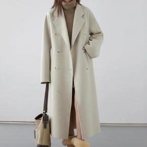 Peut inclure: Un long manteau beige clair à double boutonnage avec de grands revers. Le manteau est porté sur un pull à col roulé marron. Un sac fourre-tout beige avec des accents marron est suspendu au bras gauche. La personne porte des bottes beiges.