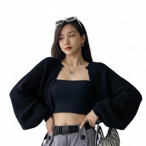 May include: A black, cropped cardigan with long, voluminous sleeves. The cardigan is paired with a black tube top, gray pants, and a black belt. A small zebra-print handbag is also visible. Sunglasses are perched on the head.