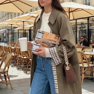 May include: A woman wearing a long olive green trench coat, blue jeans, and black sunglasses. She is holding a magazine and a white coffee cup. A small brown purse hangs from her arm. The background shows a cafe with outdoor seating.