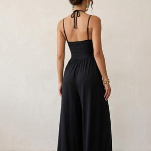 May include: A solid black jumpsuit featuring a halter neckline with thin tie straps, a fitted bodice, and wide-leg trousers. The back design includes a halter tie closure and thin shoulder straps, creating an open back silhouette with a clean finish.