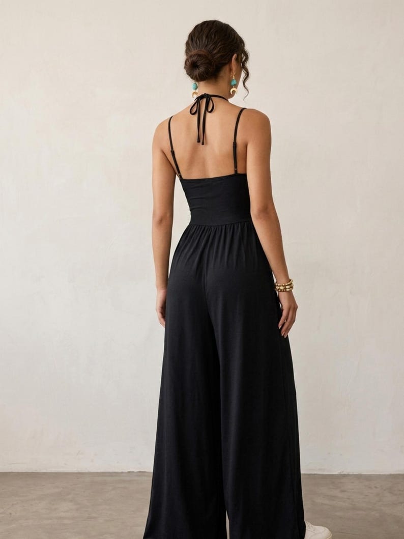 May include: A solid black jumpsuit featuring a halter neckline with thin tie straps, a fitted bodice, and wide-leg trousers. The back design includes a halter tie closure and thin shoulder straps, creating an open back silhouette with a clean finish.