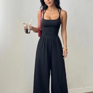 May include: A black sleeveless jumpsuit featuring a fitted bodice with a square neckline, thin shoulder straps, and a gathered waistline that transitions into wide-leg trousers. The garment is solid black and has a clean, minimalist silhouette.
