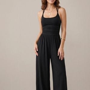 May include: A solid black jumpsuit featuring a scoop neckline, thin spaghetti straps, and a fitted bodice with subtle horizontal ruching at the waist. The garment transitions into wide-leg trousers that fall straight to the floor, creating a clean silhouette.