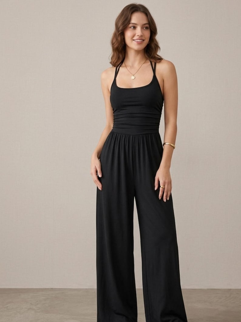 May include: A solid black jumpsuit featuring a scoop neckline, thin spaghetti straps, and a fitted bodice with subtle horizontal ruching at the waist. The garment transitions into wide-leg trousers that fall straight to the floor, creating a clean silhouette.