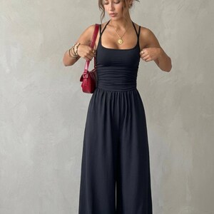 May include: A black jumpsuit featuring a fitted bodice with a square neckline, thin halter straps, and ruched detailing at the waist. The garment has wide-leg trousers and is paired with a small red shoulder bag, a gold sun pendant necklace, and stacked bracelets.