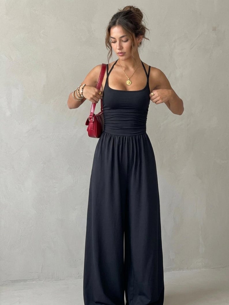 May include: A black jumpsuit featuring a fitted bodice with a square neckline, thin halter straps, and ruched detailing at the waist. The garment has wide-leg trousers and is paired with a small red shoulder bag, a gold sun pendant necklace, and stacked bracelets.