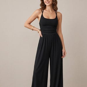 May include: A solid black sleeveless jumpsuit featuring a scoop neckline, thin spaghetti straps that cross at the back, a fitted bodice with subtle ruching at the waist, and wide-leg trousers that fall to the floor in a relaxed, flowing silhouette.