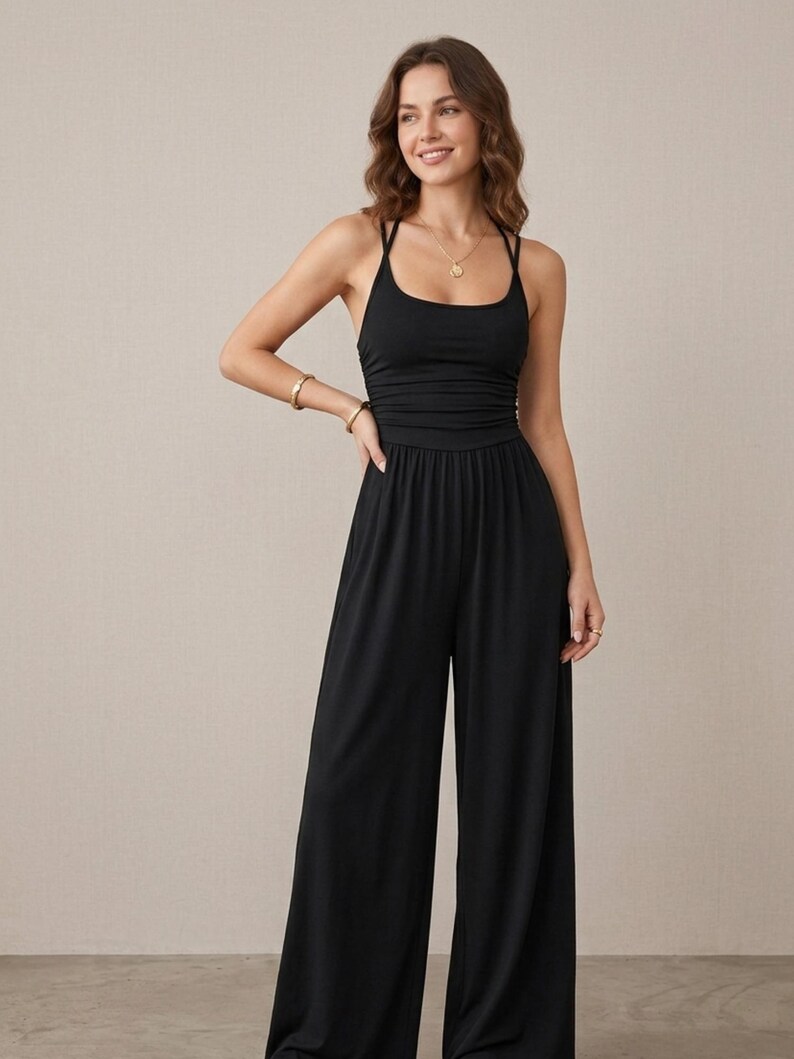 May include: A solid black sleeveless jumpsuit featuring a scoop neckline, thin spaghetti straps that cross at the back, a fitted bodice with subtle ruching at the waist, and wide-leg trousers that fall to the floor in a relaxed, flowing silhouette.