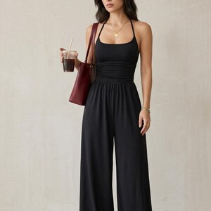 May include: A black jumpsuit featuring a halter neckline, a fitted bodice with subtle ruching at the waist, and wide-leg trousers. The garment is paired with a dark red shoulder bag and a gold pendant necklace, creating a cohesive look.
