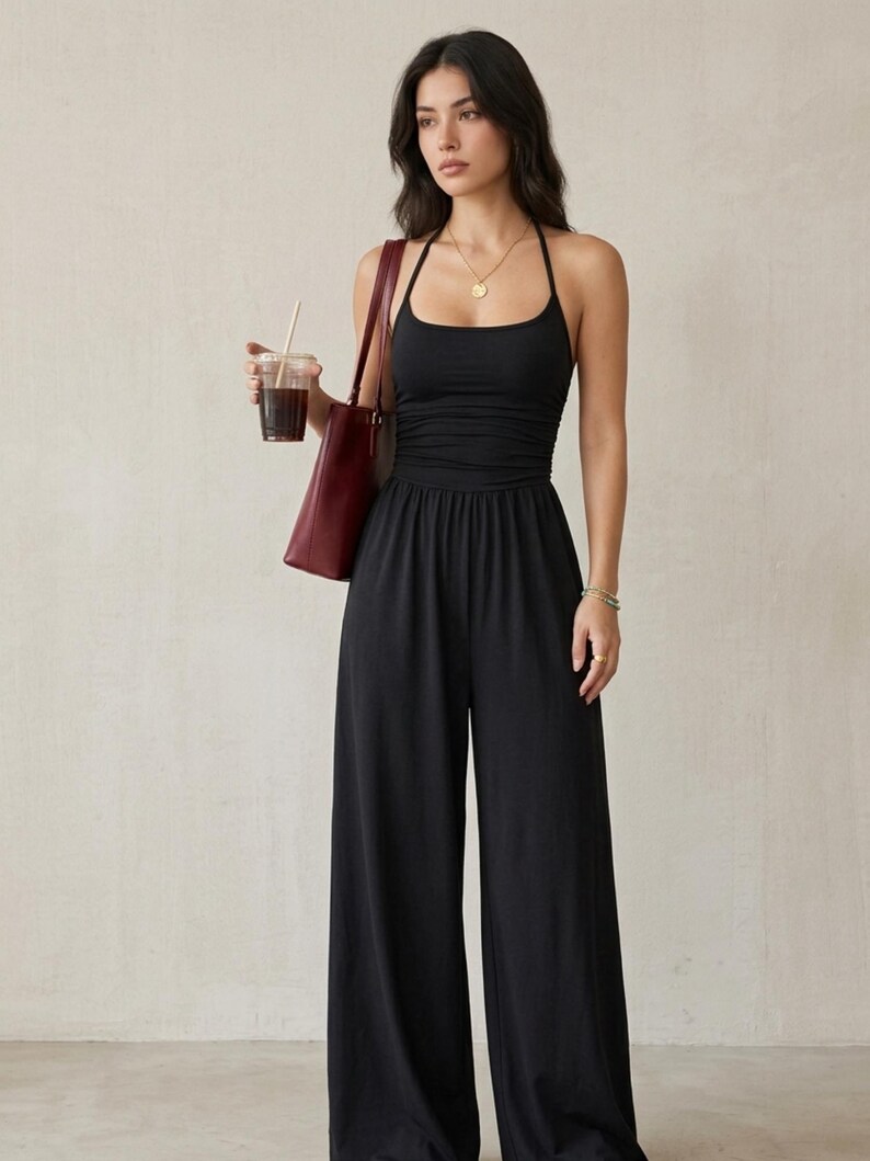 May include: A black jumpsuit featuring a halter neckline, a fitted bodice with subtle ruching at the waist, and wide-leg trousers. The garment is paired with a dark red shoulder bag and a gold pendant necklace, creating a cohesive look.