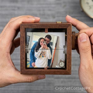 Op de afbeelding: Een vierkante houten lijst met een foto van twee personen. De lijst heeft een kleine koperen knop aan de bovenkant en een houten zwengel aan de zijkant. De foto toont een persoon in een witte hoodie met een kleurrijk patroon voor een ander.