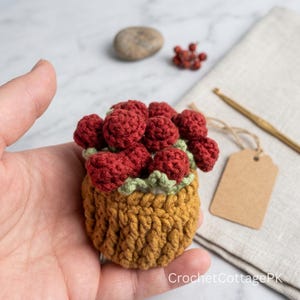 May include: A small, round, crocheted basket in mustard yellow yarn, filled with several textured, dark red crocheted berry-like shapes nestled among light green crocheted leaves. The text CrochetCottagePK is visible in the bottom right corner.
