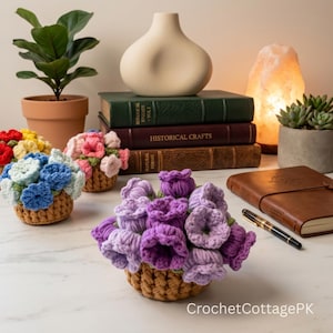 May include: A collection of three small crochet flower arrangements in tan woven baskets. One features purple and lavender flowers, another blue and white, and the third pink and yellow. The text CrochetCottagePK is visible in the bottom right corner.