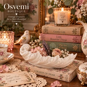 May include: A white ceramic shell-shaped bowl filled with dried flowers, resting on a book titled A Season of Home. The scene includes a candle with the text Good Things Still Happen, a floral teacup, a bird figurine, and books with floral covers.