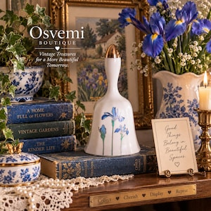 May include: A white porcelain bell with a gold-toned handle and a hand-painted blue iris floral design. The bell is positioned on a stack of vintage books, surrounded by a blue and white patterned bowl, a floral vase, and a small sign.
