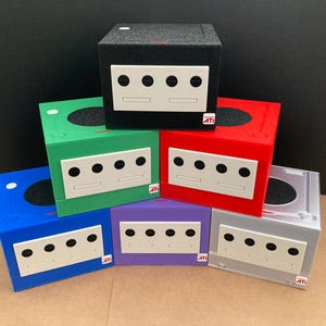 May include: A collection of six cube-shaped game consoles stacked in a pyramid formation. The consoles are black, green, red, blue, purple, and silver, each featuring a white front panel with four controller ports and a small ATI logo sticker.