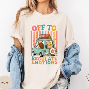 May include: An ivory t-shirt featuring a graphic of a light blue car with a person and a golden retriever inside. The car has luggage on the roof and a spare tyre on the back. The text reads OFF TO REGULATE EMOTIONS and ME TIME on the number plate.