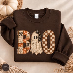 Puede incluir: Una sudadera de cuello redondo en color marrón chocolate oscuro con la palabra BOO en el pecho. La letra B es un mosaico de patrones florales y de cuadros en naranja y blanco. La letra O es un fantasma con textura de encaje y un lazo.