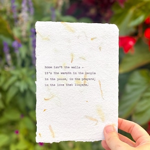May include: A handmade paper card with the quote "home isn't the walls - it's the warmth in the people..." in black text. The card has a rough, textured edge and is held against a blurred floral background.