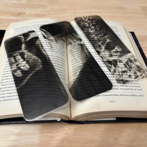 May include: Three clear, rectangular bookmarks with black and white images of a human skull and other anatomical features are placed in an open book. The bookmarks are made of transparent plastic and are resting on the pages of the book.