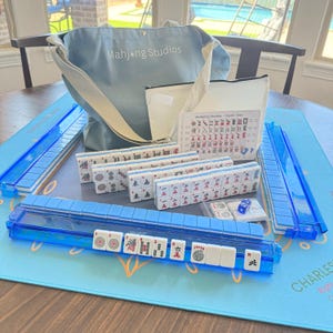 May include: A complete Mahjong set with blue accents, including tiles, dice, and a carrying bag. The set includes a blue mat and tile racks. The bag is light blue and has the text "Mahjong Studios".