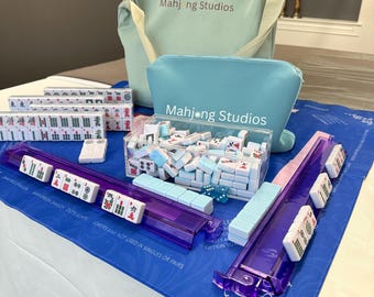 May include: A complete Mahjong set with tiles, racks, and accessories. The set includes a blue tote bag and a matching pouch, both with "Mahjong Studios" printed on them. The tiles are white with colorful symbols, and the racks are transparent purple.