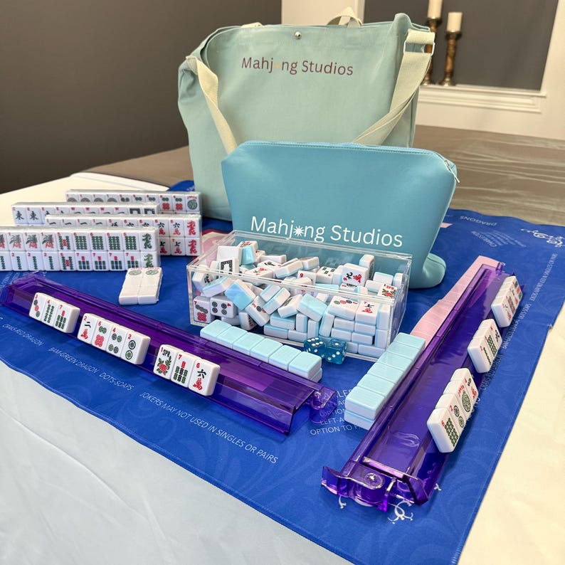 May include: A complete Mahjong set with tiles, racks, and accessories. The set includes a blue tote bag and a matching pouch, both with "Mahjong Studios" printed on them. The tiles are white with colorful symbols, and the racks are transparent purple.