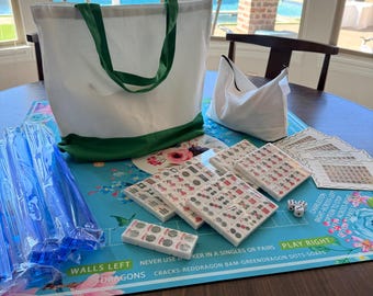 May include: A white tote bag with green handles and a green base sits on a blue floral game board. Several sets of game tiles, dice, and instruction sheets are arranged around the board. The game board has text that reads "WALLS LEFT DRAGONS".