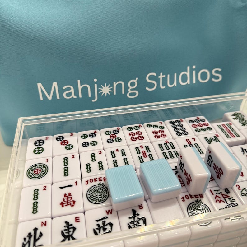 May include: A set of white Mahjong tiles with red, green, and black markings, displayed in a clear acrylic case. Several light blue tiles are visible. The text "Mahjong Studios" is printed on a light blue background.