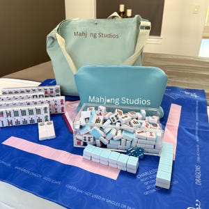 May include: A Mahjong set with a blue tote bag and pouch, both with "Mahj*ng Studios" printed on them. The set includes tiles, dice, and a blue playing mat with text. The tiles are white with red, green, and blue markings.