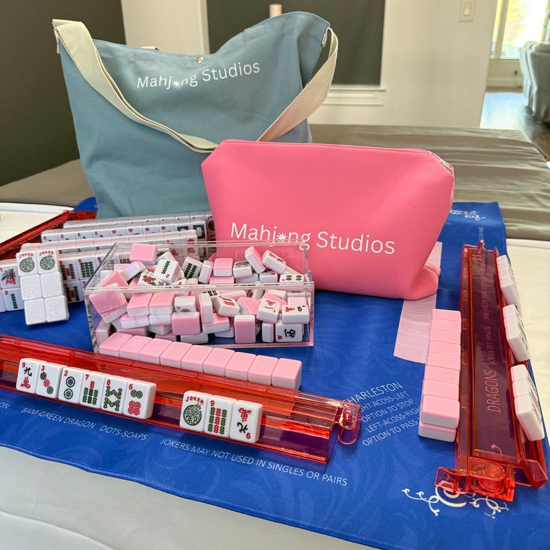 May include: A Mahjong set featuring pink and white tiles, red tile racks, and a blue tote bag. The set includes a pink zipped pouch and a clear acrylic tile holder. The text "Mahjong Studios" is printed on the bag and pouch.