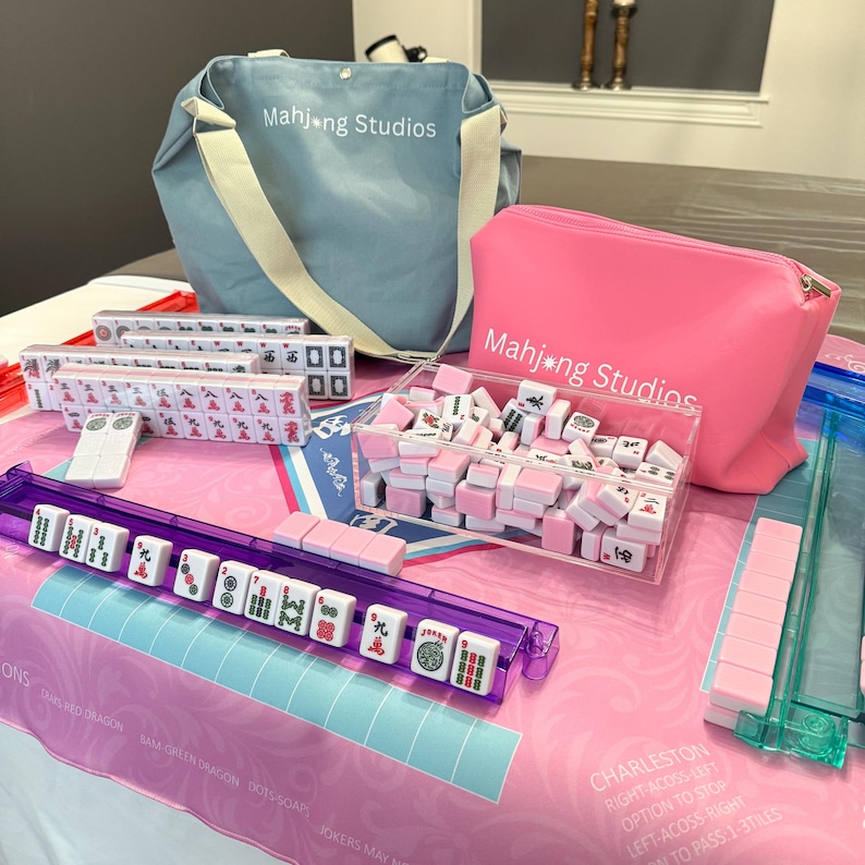 May include: A complete Mahjong set featuring pink and white tiles, a purple tile rack, and a pink carrying case. The set includes a blue tote bag and a clear acrylic box filled with tiles. The text "Mahjong Studios" is visible on the bags.