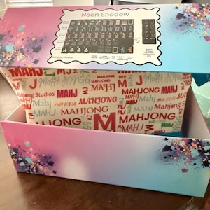 May include: A box set of the game Mahjong, featuring a colorful design with the text "Neon Shadow" on the lid. The box has a gradient of pink and blue with a patterned interior. The game tiles are visible.