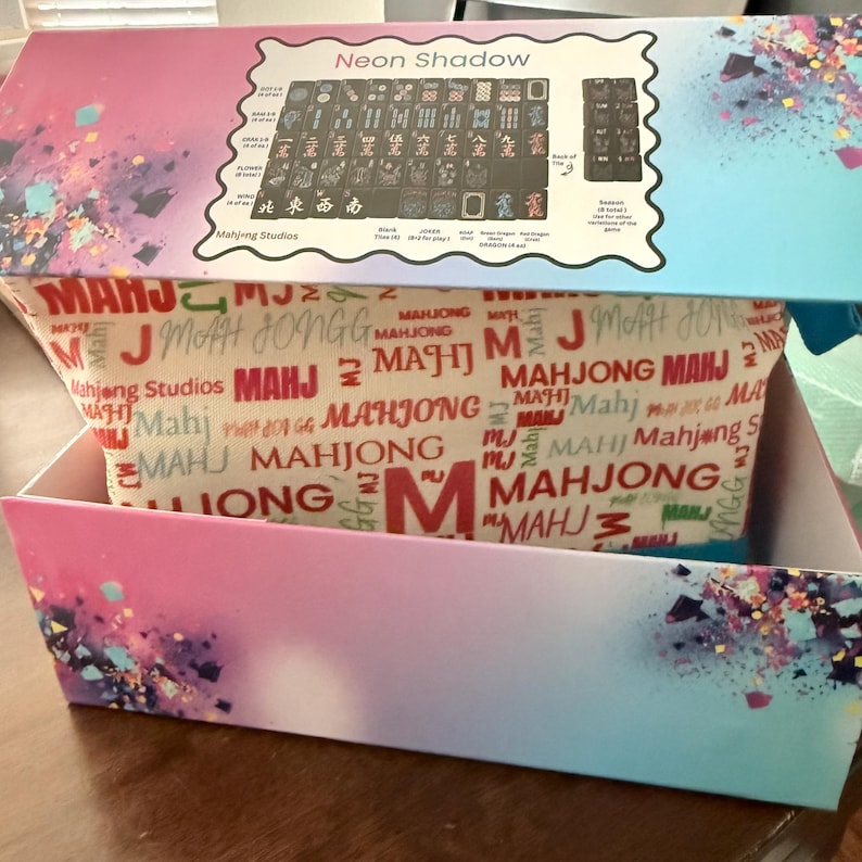May include: A box set of the game Mahjong, featuring a colorful design with the text "Neon Shadow" on the lid. The box has a gradient of pink and blue with a patterned interior. The game tiles are visible.