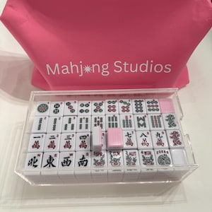 May include: A complete Mahjong set in a clear acrylic case. The white tiles feature black and red symbols and characters. A pink bag with the text "Mahj*ng Studios" is in the background.