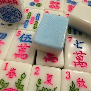 May include: Close-up of a collection of Mahjong tiles. The tiles are white with colorful symbols and numbers in pink, blue, and green. One tile has a light blue geometric pattern.