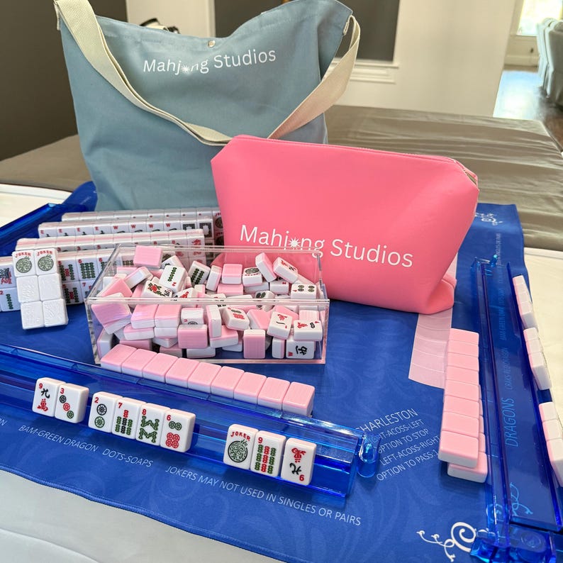May include: A Mahjong set with pink and white tiles, a clear acrylic tile holder, and blue playing mats. The set includes a light blue tote bag and a pink zippered pouch, both with "Mahjong Studios" printed on them.